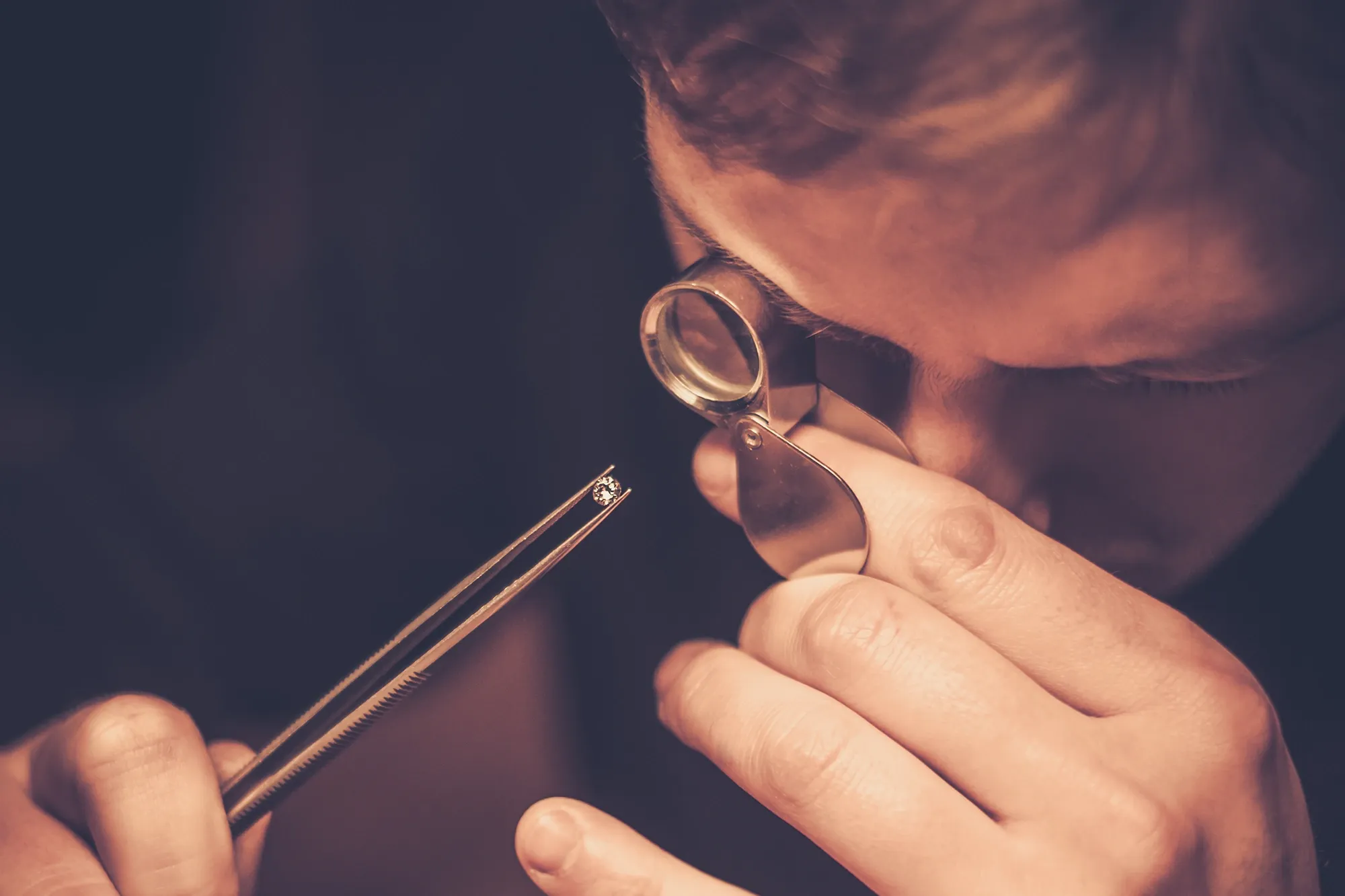 Jeweler holding a diamond in a pair of tweezers, inspecting it through a jewelers hand loupe.