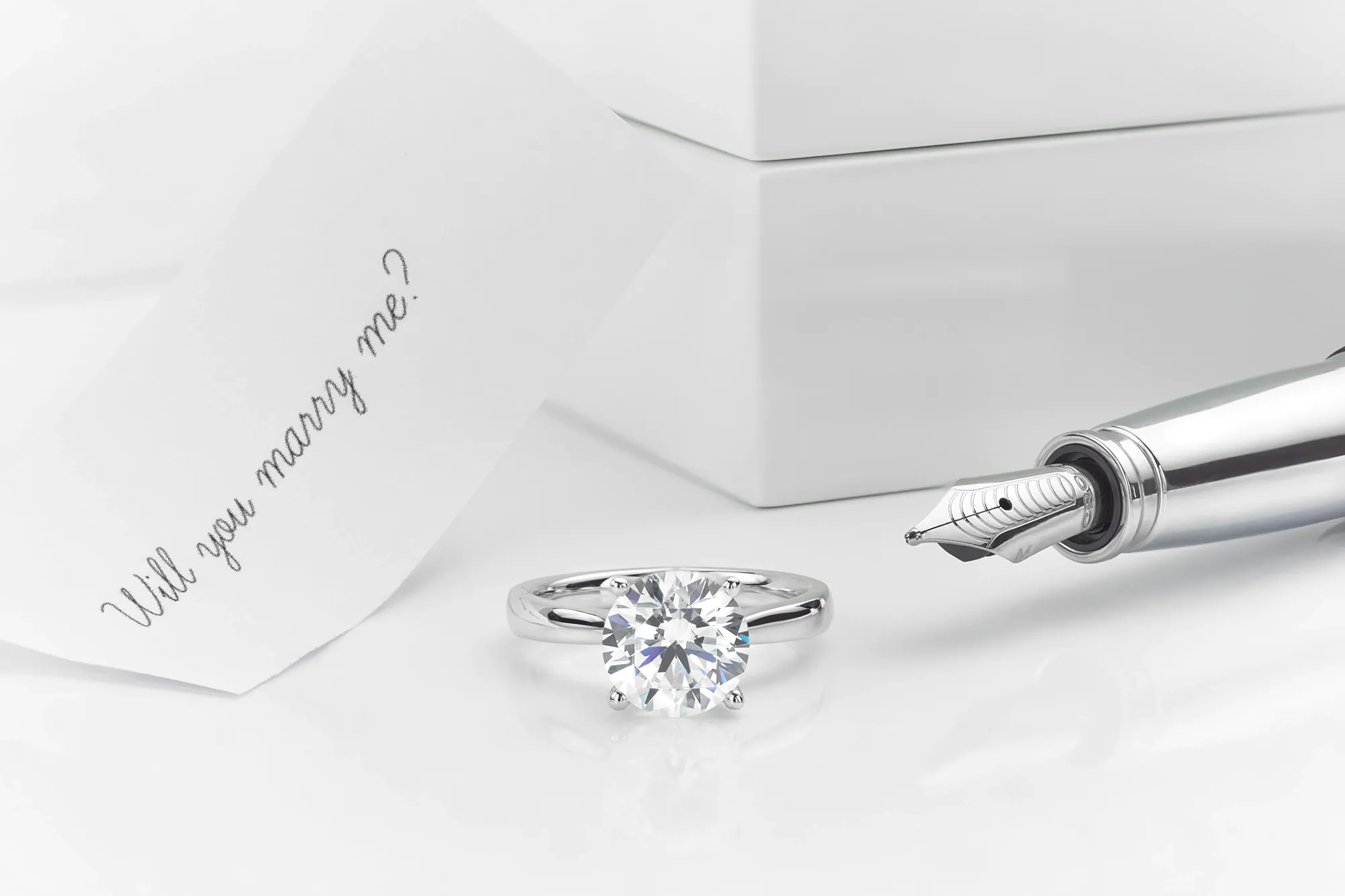 A platinum solitaire diamond ring with a fountain pen and note saying 'Will you marry me?'