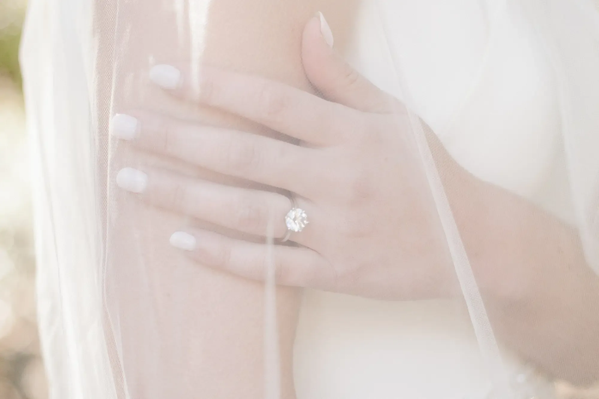 Bride's hand under her veil, wearing a round brilliant solitaire diamond ring.