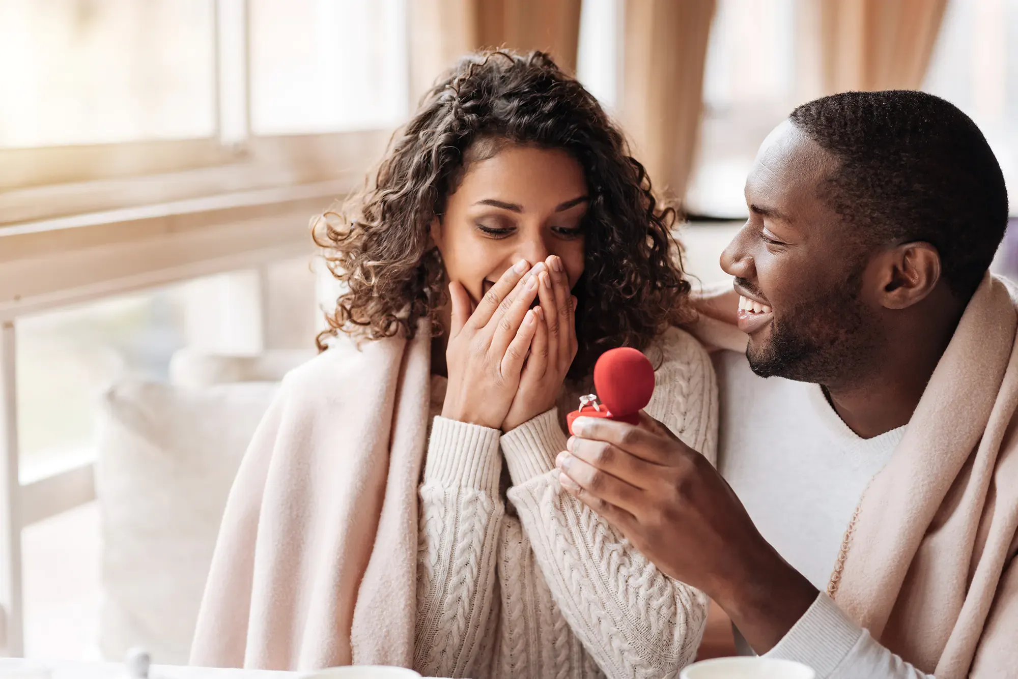 A man proposing to a smiling woman in a cafe. He is holding an open red ring box with engagement ring.