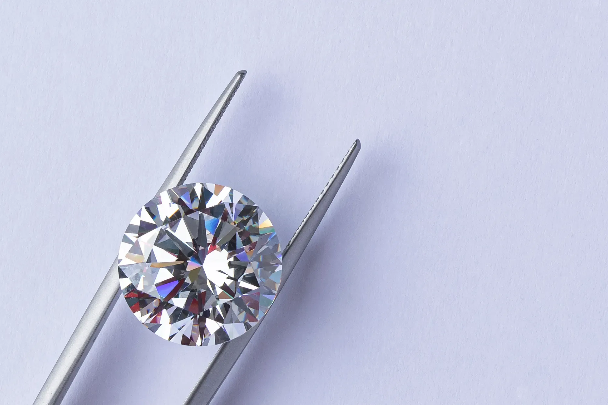 A round brilliant cut diamond in tweezers set against a purple background.