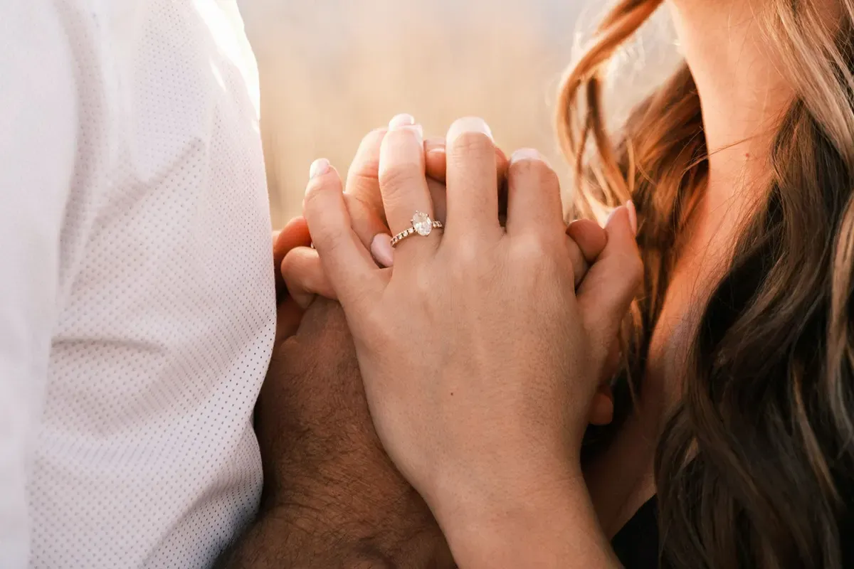 A couple standing close together, hands lovingly intertwined. The woman's hand shows a pavé diamond engagement ring with an oval diamond center stone.