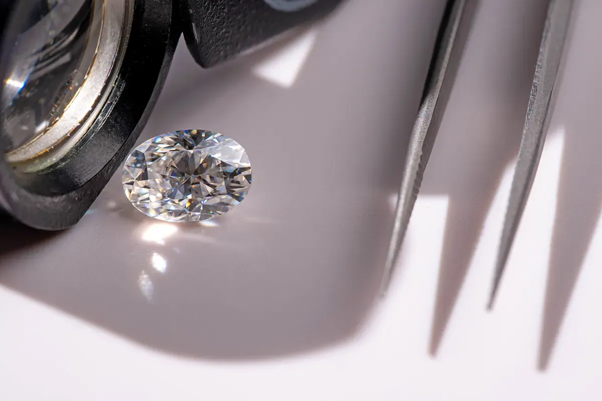 A loose oval cut diamond sparkles in the light. It sits next to a jeweler's loupe and a pair of tweezers.