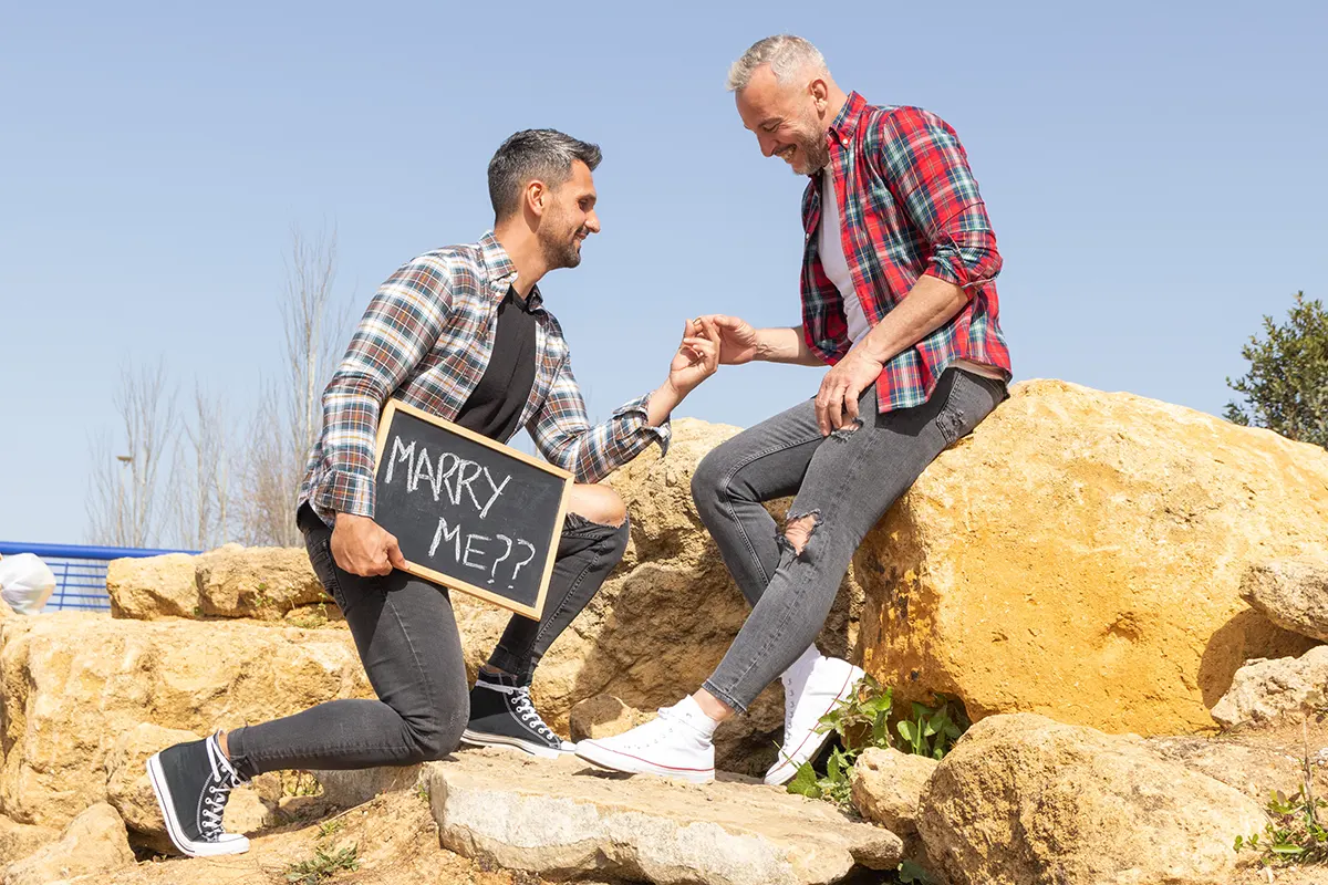 A male gay couple posing on boulders. One is down on one knee proposing marriage with a chalk board saying 'Will you Marry Me?'